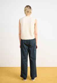 Pulover fără mâneci, tricotat, de culoare crem, cu un model în cablu, asortat cu pantaloni negri, largi. Fundalul prezintă un podea bej și un perete simplu.