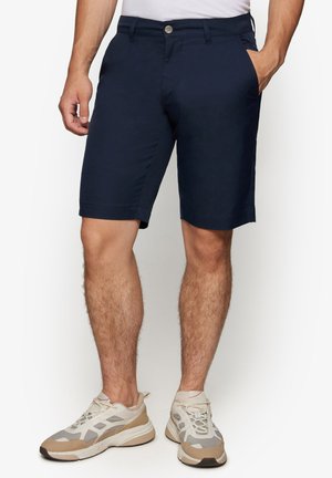Mand iført marineblå afslappede shorts og beige atletiksneakers, stående med den ene hånd i lommen mod en hvid baggrund.