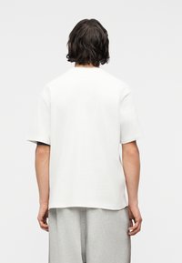 Άτομο με μακριά μέχρι τους ώμους σκουρόχρωμα μαλλιά, που φοράει απλό λευκό κοντομάνικο πουκάμισο και ανοιχτό γκρι παντελόνι, γυρισμένο πλάτη σε απλό φόντο.