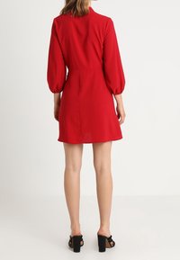 Červené šaty z hladkého materiálu, s balónovými rukávy, přiléhavým tělem a rozšířeným okrajem. Nosené s černými botami na blokovém podpatku.