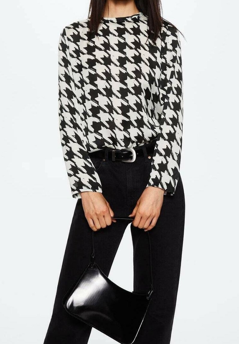 Femme portant un haut à manches longues pied-de-poule noir et blanc et un pantalon noir, tenant un petit sac à main noir par la poignée.
