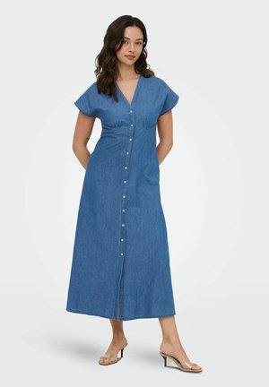 Donna con capelli scuri che indossa un vestito lungo blu con bottoni e maniche corte, e sandali con tacco trasparente, in piedi con le mani dietro la schiena.