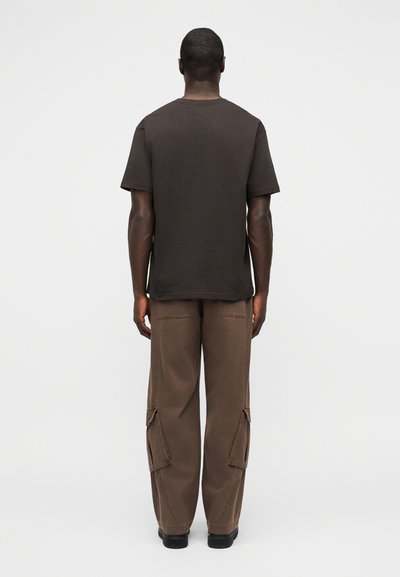 Άνδρας με απλό μαύρο κοντομάνικο μπλουζάκι και φαρδιά καφέ παντελόνια cargo, στέκεται με την πλάτη προς τα εμπρός σε απλό φωτεινό φόντο.