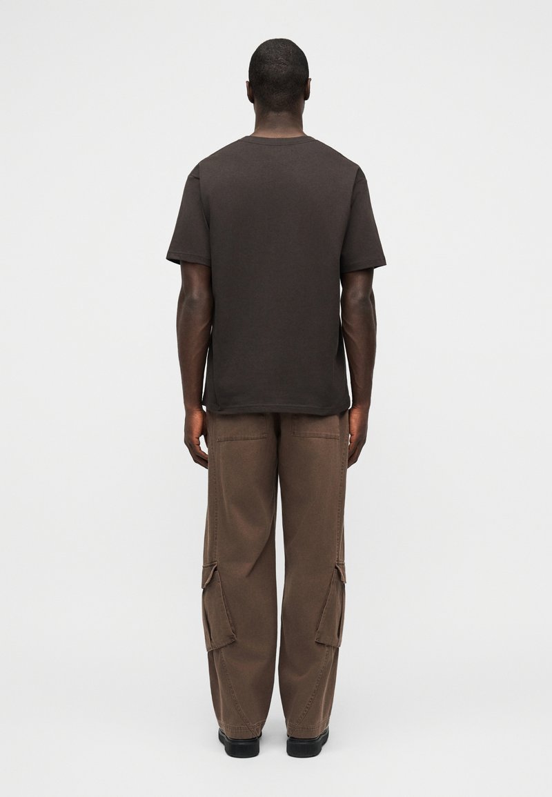 Man wearing a plain black short-sleeve T-shirt and loose brown cargo pants, standing facing away on a plain light background.