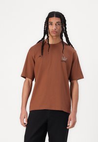 Browns kokvilnas t-krekls ar īsām piedurknēm un apaļu kakla izgriezumu; uz krūšu daļas ir neliels melns Adidas logo. Gluda tekstūra.