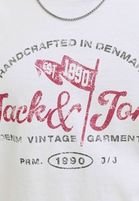 Balti medvilninė marškinėliai su raudonu ir juodu grafikiniu tekstu, vėliavomis ir 1990 metų numeriu, su vintage stiliaus spauda ir sidabriniu grandininiu kaklo papuošalu.