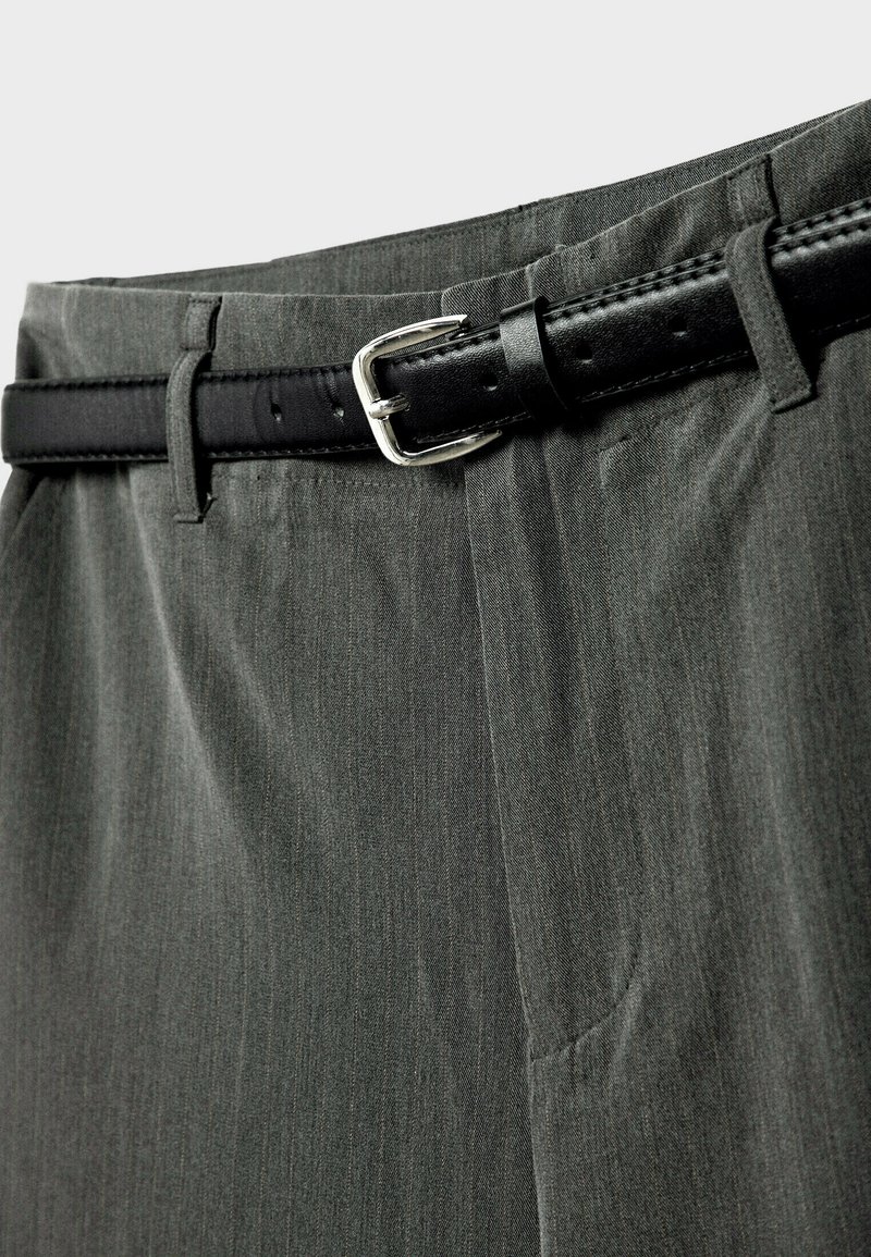 Gros plan sur un pantalon de costume gris maintenu par une ceinture en cuir noire avec une boucle en métal argenté.