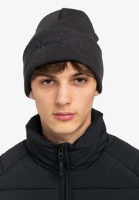 Beanie grigio lavorato a maglia con una texture a coste e un logo ricamato di blu scuro. Indossato con una giacca nera con zip e colletto alto.