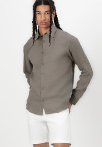 Camisa de lino verde oliva con cuello, cierre frontal de botones y mangas largas, acompañada de pantalones cortos blancos. Textura suave, diseño informal.