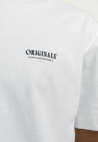 T-shirt bianca in cotone con un logo stampato nero "ORIGINALS™" e "© DAL DICANNOVE NOVANTA" sottostante, leggermente curvato per evidenza.