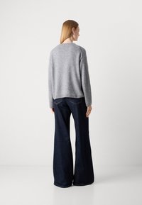 Mulher de costas, vestindo um suéter cinza e jeans largos escuros, contra um fundo branco liso.