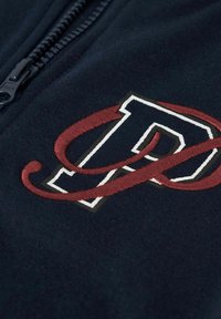 Granatowy rozpinany sweter z dużym haftowanym logo w czerwono-białych kolorach, umieszczonym na tle teksturowanej tkaniny.