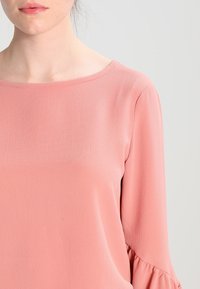 Blouse couleur pêche avec une texture lisse, encolure ronde et manches longues ornées de subtils volants aux poignets.