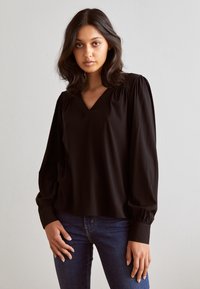 Blusa nera con scollo a V e design con maniche a sbuffo. Tessuto liscio, vestibilità comoda, dettagli arricciati sulle spalle, abbinata a jeans scuri.