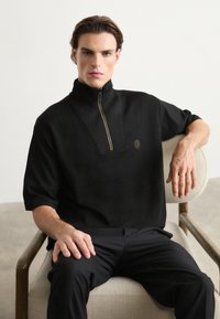 Juodas zip-neck puloveris su tekstūruotu paviršiumi, trumpomis rankovėmis, auksiniu užtrauktuku ir apvaliu logotipo akcentu ant krūtinės.