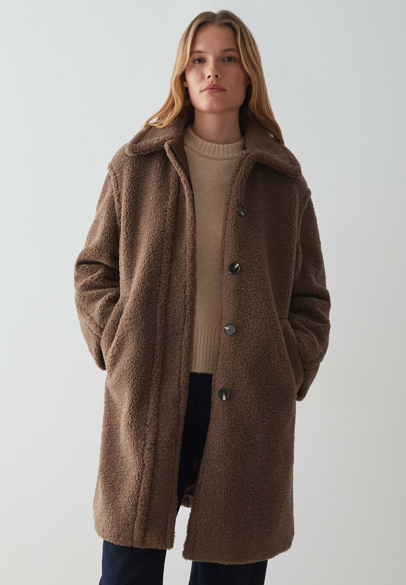 Manteau en polaire marron avec un col, des boutons sur le devant et des poches latérales ; texture douce ; associé à un pull beige clair en dessous.