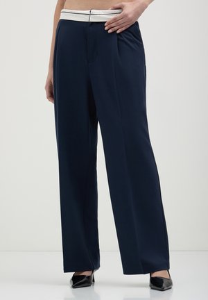 Pantalons larges bleu marine en tissu lisse, avec une taille bande blanche et des plis à l'avant, associés à des chaussures noires à bout pointu.