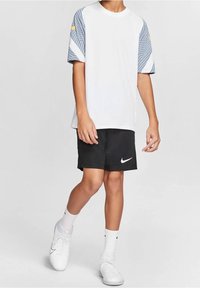 Vit t-shirt med mönstrade grå ärmar, svarta shorts med vit Nike-logotyp och vita skor. Materialet verkar lätt och sportigt.