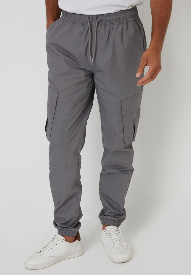 Homme portant un pantalon cargo jogger gris avec des poignets élastiques et des poches, associé à des baskets blanches.