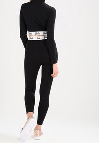 Svart långärmad bodysuit med hög hals och vit elastisk midja med "Barbie"-logotyp, kombinerad med leggings och skor i roséguld.