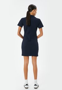 Granatowa sukienka polo z krótkim rękawem, o prostym kroju, z dwoma bocznymi kieszeniami i szwem na plecach dla podkreślenia stylu. W zestawie z białymi trampkami i skarpetkami.