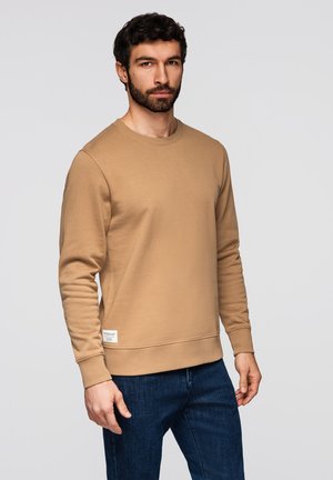 Man met donker krullend haar en baard, gekleed in een beige lange-mouwen trui en donkerblauwe spijkerbroek, tegen een effen achtergrond.