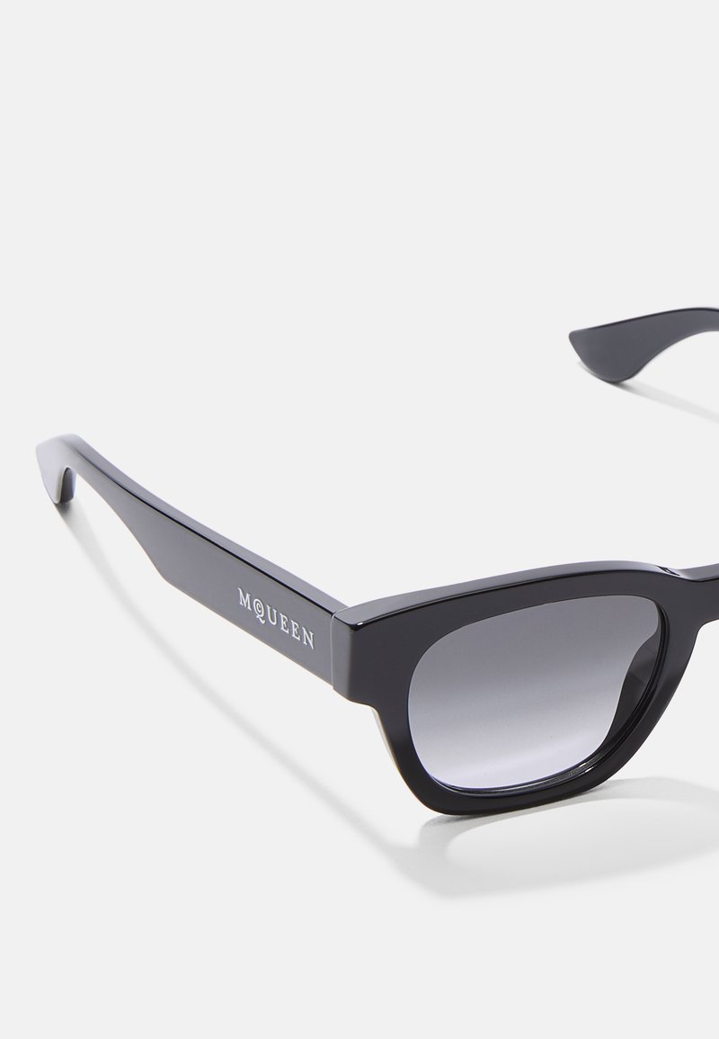 Schwarze Sonnenbrille mit katzenaugenförmigem Design und grauen Verlaufsgläsern. Das Gestell hat eine glänzende Oberfläche und das "MQUEEN"-Logo an der Seite.