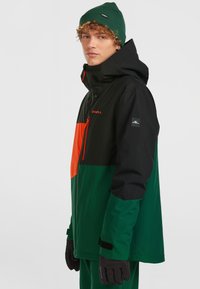 Groene, zwarte en oranje skijack met een ritssluiting aan de voorkant, kap en logo. Beschikt over een kleurblokontwerp en handschoenen. Lichtgewicht stof.
