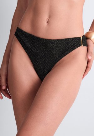 Femme portant un bas de bikini noir texturé avec des détails de boucle dorée sur les hanches sur un fond uni.