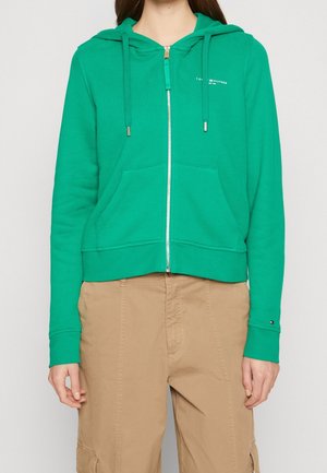Frau trägt einen grünen Hoodie mit Reißverschluss und Fronttaschen sowie beige Cargohosen und steht vor einem schlichten hellen Hintergrund.