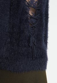 Marineblauer, flauschiger Pullover mit einem Spitzenmuster darunter. Verfügt über eine Schnürdetail vorne und eine weiche, strukturierte Stoffoberfläche.