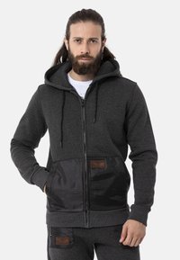 Zip-up hoodie in dark gray fabric with textured finish. Features a black satin front panel and two pockets with leather accents.