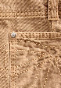 Tissu en denim beige avec un accent de matériel argenté en forme d'étoile. Présente des coutures visibles et un motif unique autour de la zone de la poche.