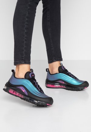 Persoon die zwarte skinny jeans en zwarte, blauwe en roze Nike Air Max sneakers draagt, staand op een witte vloer.