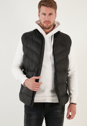 Schwarze Steppweste mit Chevron-Design, Reißverschluss vorne und Logodetail. Über einem weißen Hoodie getragen, mit glatter, glänzender Textur.