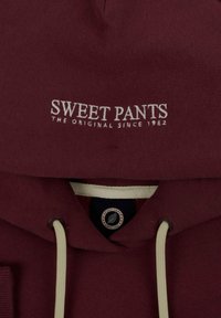 Hoodie bordeaux avec une poche avant, des cordons de crème et un texte brodé blanc indiquant "SWEET PANTS L'ORIGINAL DEPUIS 1982."