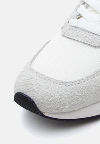 Jasnoszary but sportowy z fakturowanym zamszowym czubkiem, siateczkową cholewką, białymi sznurowadłami i czarną gumową podeszwą.