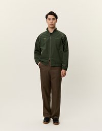 Zelená bunda na zips s elastickými manžetami a jemnými švovými detailmi, skombinovaná s hnedými kockovanými nohavicami a čiernymi topánkami.