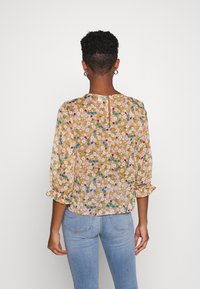 Frau mit kurzem lockigem Haar, die eine senfgelbe Bluse mit Blumenmuster und ballonförmigen Ärmeln sowie eine hellblaue Jeans trägt, von hinten gezeigt.