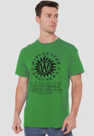 Man wearing green T-shirt with black "Winchester Brothers" design and text "Saving people hunting things, Supernatural, Join the hunt."