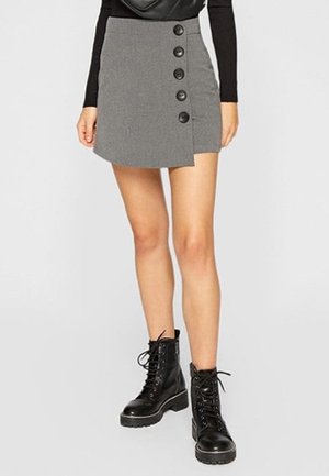 Vrouw draagt een grijze minirok met grote knopen aan de zijkant, een zwarte top met lange mouwen en zwarte veterboots met dikke zolen.