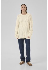 Frau, die in einem cremfarbenen, grob gestrickten Oversize-Pullover, dunkelblauen Jeans und braunen Sneakers vor einem schlichten grauen Hintergrund steht.