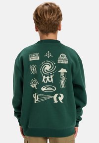 Tmavohnedá mikina so svetlými grafickými potlačami na chrbte, vrátane log a abstraktných vzorov. Mäkky materiál.