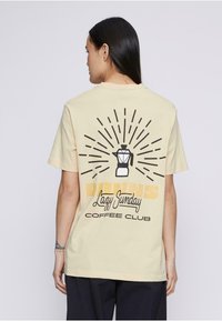 Camiseta beige claro con un gráfico en la parte posterior que presenta una cafetera y el texto: "Club de Café del Domingo Perezoso". Mangas cortas y corte relajado.