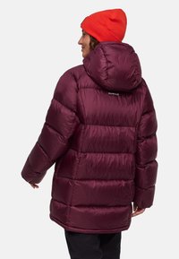 Maroen pufferjack met een capuchon, gequilt ontwerp en glanzende textuur. Een rode gebreide beanie wordt op het hoofd gedragen, met zichtbaar merklogo op de achterkant.