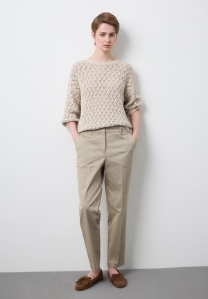 Woman standing against white wall wearing beige knitted sweater, khaki trousers, and brown moccasin shoes with hands in pockets.