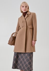 Brauner Wollmantel mit maßgeschneidertem Design, zweireihigen Knöpfen, Reverskragen und Seitentaschen, kombiniert mit einem karierten Rock und einer braunen Handtasche.