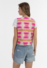 Colorful woven vest with pink and orange patterns, worn over a white shirt, paired with blue denim shorts. Textured fabric, cropped design.