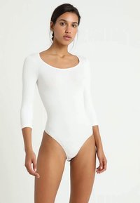 Nainen, joka on pukeutunut tiiviiseen valkoiseen pitkähiharaiseen bodysuitsiin, seisoo yksinkertaista valkoista taustaa vasten neutraalilla ilmeellä.