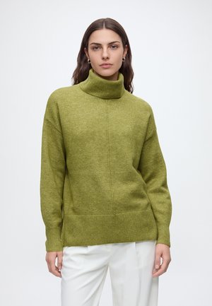 Femme aux longs cheveux foncés portant des boucles d'oreilles anneaux dorés, un pull à col roulé vert olive et un pantalon blanc, debout devant un fond uni.